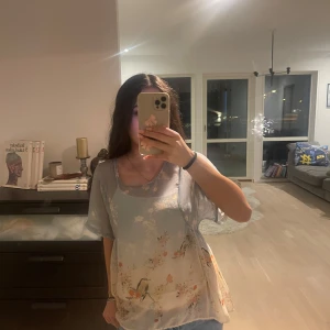 Blommig blus från Vintage - Säljer en luftig blommig blus från Vintage i ljusblått med korta ärmar. Blusen har ett vackert blommönster i rosa och beige toner, perfekt för en somrig look. Den är tillverkad i ett lätt och skönt material.