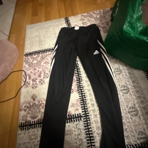 Svarta träningsbyxor från Adidas - Säljer ett par svarta träningsbyxor från Adidas med vita detaljer och logga på sidan. Byxorna har en elastisk midja och är perfekta för sport och träning. De är tillverkade i ett lätt och bekvämt material.