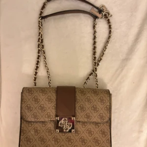 Beige axelväska med kedjedetaljer - Snygg beige axelväska med mönster och kedjedetaljer på axelremmen. Väskan har en elegant design med ett metallspänne framtill och en brun remdetalj. Äkta Guess väska   Knappt använd