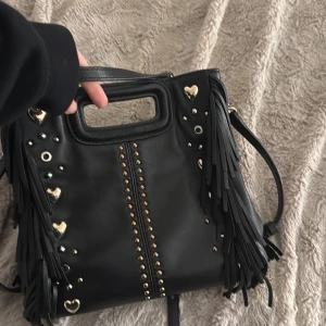 Maje Väska - Min fina snygga maje väska! Använd men i så bra skick! Har dustbagen och långt band till som tillkom! Snygg svart axelväska i skinn med fransar och dekorativa hjärtan och nitar. Väskan har ett praktiskt handtag och en justerbar axelrem. Perfekt för att ge din outfit!