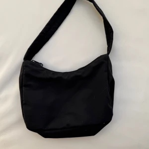 Svart axelväska från Gina Tricot - Snygg och praktisk svart axelväska från Gina Tricot. Väskan har en dragkedja upptill och en justerbar axelrem. Perfekt för att bära dina dagliga nödvändigheter med stil.