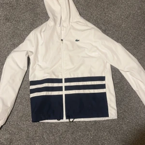 Vit och marinblå vindjacka och byxor från Lacoste - Snygg vit vindjacka och byxor från Lacoste med marinblå detaljer och dragkedja. Jackan har en huva och elastiska muddar. Perfekt för en sportig look.
