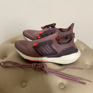 Adidas Ultraboost i lila och orange - Säljer ett par Adidas Ultraboost i en snygg lila nyans med orange detaljer. Skorna har en bekväm och responsiv sula, perfekt för löpning eller vardagsbruk. De är tillverkade i syntetmaterial och har en stilren design utan snörning.