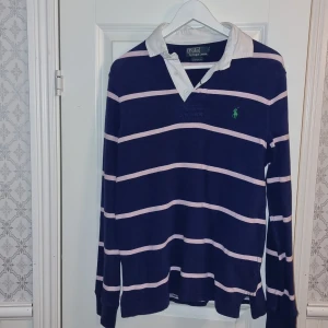 Randig pikétröja från Ralph Lauren - Snygg randig pikétröja från Ralph Lauren i blått och rosa. Tröjan har långa ärmar och en klassisk krage med knappar. Perfekt för en stilren och avslappnad look.