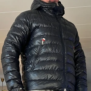 Svart dunjacka från Moncler - Säljer en snygg svart dunjacka från Moncler med dragkedja och huva. Jackan har en quiltad design och praktiska fickor med dragkedjor. Perfekt för kyliga dagar. 2 st små lagningar finns , en på insidan nertill på ryggen , samt en nertill på framsidan , skickar gärna bilder på dessa om så önskas .. Jackan passar mig som använder medium .. 