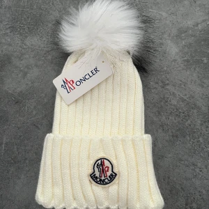 Vit stickad mössa från Moncler - Snygg vit stickad mössa från med en fluffig pälsboll på toppen. Mössan har ett ribbat mönster och Monclers logotyp framtill. Perfekt för kyliga dagar!