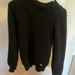 Svart ribbad tröja🖤 - Säljer en svart ribbad tröja med långa ärmar och rund hals. Tröjan har en unik textur och en liten metallbricka vid nederkanten. Perfekt för kyliga dagar. Från ”Dobber since 1974” i storlek XS. Använd & i mycket bra skick😊