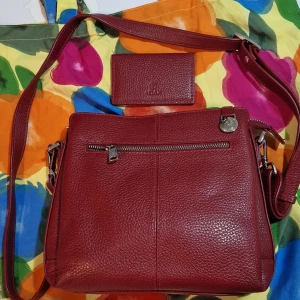 Röd axelväska från Adam - Snygg röd axelväska i äkta skinn från Adax med justerbar axelrem och praktiska fack. Väskan har en dragkedja upptill och en extra ficka med dragkedja på framsidan. Perfekt för att hålla ordning på dina saker.