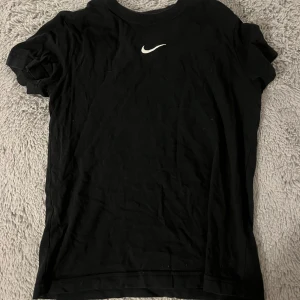 Svart t-shirt från Nike - Säljer en svart t-shirt från Nike med deras ikoniska logga på bröstet. T-shirten har en loose fit och är tillverkad i bomull, perfekt för en avslappnad stil och även för träning. Aldrig använd