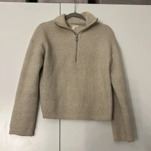 Beige stickad tröja med dragkedja - Säljer en stilren beige stickad tröja från H&M. Tröjan har en halv dragkedja och en hög krage, perfekt för kyliga dagar. Den är ribbad och har långa ärmar, vilket ger en klassisk look. Passar utmärkt till både vardag och mer avslappnade tillfällen.