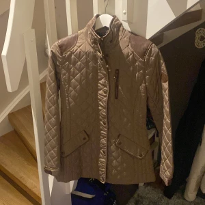 Beige quiltad jacka från Ralph Lauren - Snygg beige quiltad jacka från Ralph Lauren med bruna axelpartier och detaljer. Jackan har en hög krage och dragkedja framtill samt praktiska fickor med knappar. 
