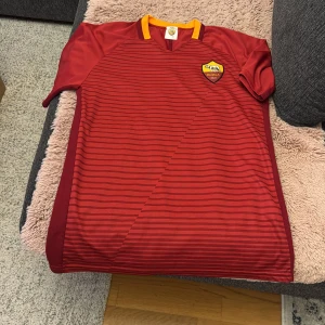 Röd AS Roma fotbollströja - Säljer en röd AS Roma fotbollströja med gula detaljer och klubbens emblem på bröstet. Tröjan har korta ärmar och ett randigt mönster. Namnet Džeko och nummer 9 är tryckt på baksidan.