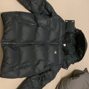 Blå dunjacka från Calvin Klein - Snygg blå dunjacka från Calvin Klein med dragkedja och avtagbar huva. Perfekt för kalla dagar med sin pufferdesign som håller dig varm. Jackan har två praktiska fickor med dragkedja framtill.
