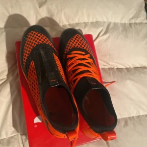 Orange och svarta fotbollsskor från Puma - Snygga fotbollsskor från Puma i orange och svart med ett unikt nätmönster. Skorna har snörning och är designade för smal passform. Perfekta för den som vill sticka ut på planen. Använda endast en gång