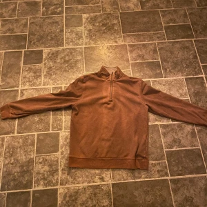 Brun halfzip tröja från Van Heusen - Säljer en brun halpzip tröja från Van Heusen med dragkedja vid halsen. Perfekt för kyliga dagar med sin mjuka och varma insida. Tröjan har långa ärmar och en klassisk passform.