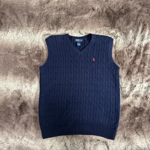 Polo Ralph Lauren - Säljer en snygg mörkblå stickad väst från Polo Ralph Lauren med v-ringning och ett rött broderat emblem på bröstet. Perfekt för lager-på-lager under höst och vår.