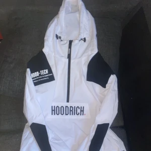 Hoodrich windbreaker  - Snygg vit vindjacka från Hoodrich med svart text och detaljer. Jackan har en dragkedja framtill och en justerbar huva. Perfekt för blåsiga dagar.