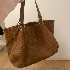 Brun handväska  - Snygg brun axelväska med flätat mönster och dubbla handtag. Tyvärr har den blivit lite sliten högst upp vid handtagen som syns på bilden men annars är den i jättebra skick 