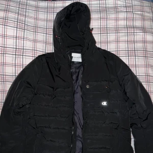 Svart dunjacka från Calvin Klein - Snygg svart dunjacka från Calvin Klein med huva och dragkedja. Jackan har en diskret logga på bröstet och praktiska fickor med knappar. Perfekt för kyliga dagar.endast andvänt 2 gånger. 