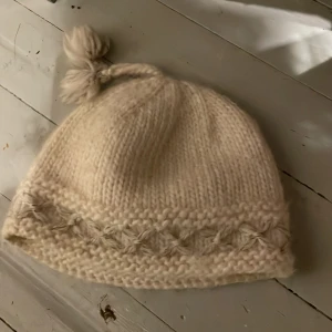 Ull mössa - Mysig beige stickad mössa med en dekorativ tofs på toppen. Den har ett fint mönster längs kanten som ger extra stil. Perfekt för kyliga dagar! Ull
