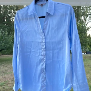 Blå skjorta i satin - En ljusblå skjorta i satin från H&M. Köptes ett par år tillbaka men blev dessvärre aldrig använd 🩵 Superskön och tunn i materialet. Passar bra för sig själv eller som overshirt över en topp exempelvis 😚  (ALDRIG ANVÄND)  Längd: 68 cm Axel: 40 cm