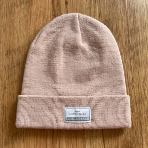 Rosa mössa från 157 Denim Makers - Säljer en superfin rosa mössa från 157 Denim Makers. Den är gjord i 100% akryl och har en stilren design med en liten etikett framtill. Perfekt för att hålla sig varm och snygg under kyliga dagar! I ny skick, inga hål eller fläckar. 