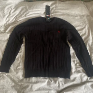 Kabelstickad tröja - Hej! Jag säljer denna kabelstickade tröja som jag enbart använt en gång men inte föll mig i smaken. Den är så gott som ny, den är svart och har ett litet rött emblem med en gubbe på en häst. Storlek m och sitter så den passar både S och m. 