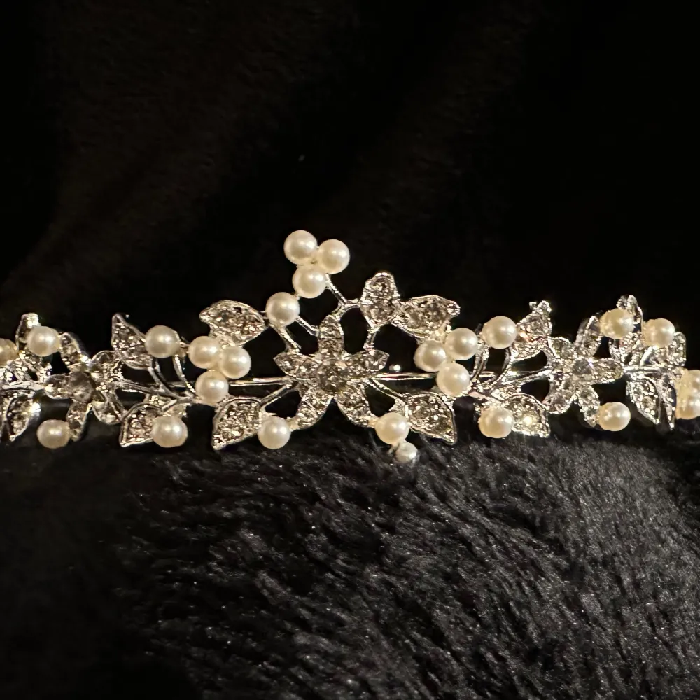 Säljer en supervacker tiara som är perfekt för speciella tillfällen. Det är dekorerat med glittrande strass och eleganta pärlor som ger en lyxig känsla. Tiaran har en blommig design som gör det extra romantiskt och stilfullt. Höjd 3 cm längst fram.. Accessoarer.