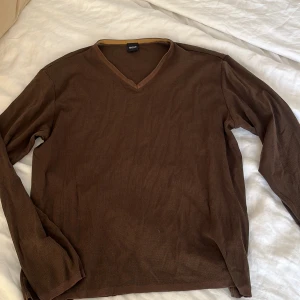 Brun tröja från Hugo Boss - Säljer en snygg brun tröja från Hugo Boss i storlek L. Den är gjord av 54% bomull och 46% nylon, vilket gör den både bekväm och hållbar. Tröjan har en stilren v-ringning och långa ärmar, perfekt för både vardag och lite finare tillfällen. Passar bra till höst och vår!