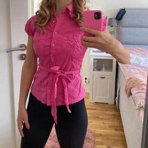 Rosa blus med knytband - Säljer en superfin rosa blus med korta ärmar och knytband i midjan. Blusen har knappar framtill och två bröstfickor. Perfekt för sommardagar eller en festlig kväll! 💖