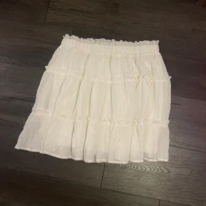 Vit kjol från Shein - Säljer en söt vit kjol från Shein med volangdetaljer. Kjolen har en elastisk midja och är perfekt för en avslappnad stil. Den är luftig, vilket gör den idealisk för varmare dagar.