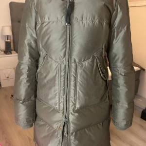 Grön dunjacka från Parajumper - Snygg och varm grön dunjacka med dragkedja och pälsdetalj på huvan från parajumper. Perfekt för kalla dagar med sin långa design och praktiska fickor. Säljes ensast för att den är för liten 