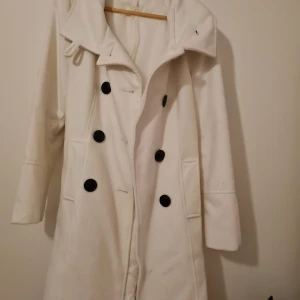 Vit kappa med svarta knappar - Elegant vit kappa med stora svarta knappar och bälte i midjan. Perfekt för kyliga dagar med sin långa design och stilrena look. Kappan har långa ärmar och en klassisk krage.