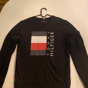 Svart tröja från Tommy Hilfiger - Snygg svart tröja från Tommy Hilfiger med det ikoniska logotyptrycket i rött, vitt och blått på framsidan. Tröjan har långa ärmar och en klassisk rund halsringning. Perfekt för en stilren och avslappnad look.svart färg 