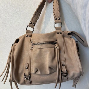 Beige axelväska med fransar - Snygg beige axelväska med flätade handtag och fransdetaljer. Väskan har flera dragkedjor och nitar för en cool look. Perfekt för att bära med sig det nödvändigaste med stil.