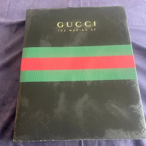 Gucci: The Making Of - Säljer boken 'Gucci: The Making Of' med ett elegant svart omslag prytt av det ikoniska gröna och röda bandet. Perfekt för modeentusiaster och Gucci-fans som vill fördjupa sig i varumärkets historia och design.