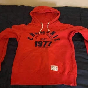 Röd hoodie från Cover - Säljer en snygg röd hoodie från Cover med tryck 'Chamonix 1977' på framsidan. Den har en känguruficka och justerbar huva med vita snören. Perfekt för en avslappnad stil.
