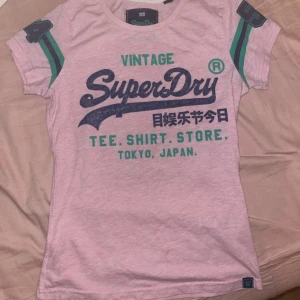 Rosa t-shirt från SuperDry - Snygg rosa t-shirt från SuperDry med tryck i blått och grönt. Den har korta ärmar med gröna och blå detaljer. Perfekt för en avslappnad stil.