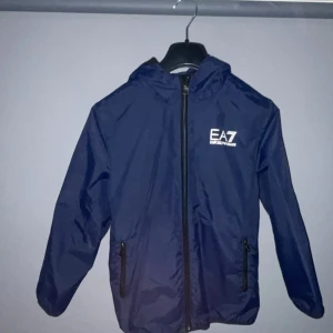Blå vindjacka från Emporio Armani - Snygg blå vindjacka från Emporio Armani EA7 med huva och dragkedja. Perfekt för blåsiga dagar. Jackan har en stilren design med logotyp på bröstet. Den passar folk som är mellan 153-156. Diskutera gärna pris med mig.