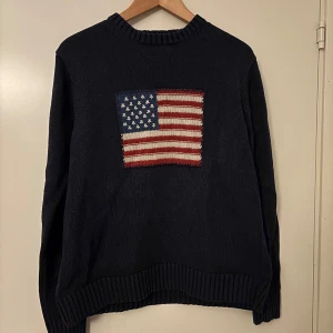 Mörkblå tröja från Gant - Säljer en mörkblå tröja från Gant med en stor amerikansk flagga på framsidan. Tröjan är tillverkad i 100% bomull och har ribbade kanter vid hals, ärmslut och nederkant. Perfekt för en casual look med en touch av amerikansk stil.