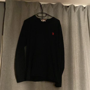 Svart tröja från U.S. Polo Assn. - Snygg svart tröja från U.S. Polo Assn. med ett rött broderat logotypmotiv på bröstet. Tröjan har långa ärmar och en klassisk passform. Perfekt för en bakotlutat still och storleken är M men passar lika bra på S