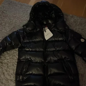 Svart pufferjacka från Moncler - Säljer en snygg svart pufferjacka från Moncler som är helt ny. Jackan har en glansig finish och är utrustad med dragkedja och huva. Perfekt för kyliga dagar när du vill hålla stilen. 🖤