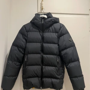 Svart dunjacka från J.Lindeberg - Snygg svart dunjacka från J.Lindeberg med huva och dragkedja. Jackan har en quiltad design och ett gult foder som ger en cool kontrast. OANVÄND! Storlek är XS, passar även S. Herrmodell.