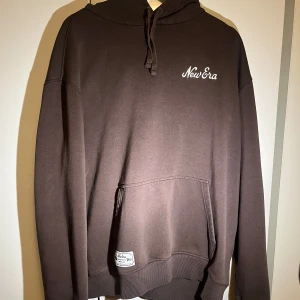 Brun hoodie från New Era - Säljer en brun hoodie från New Era med en klassisk design. Hoodien har en känguruficka och justerbar huva med dragsko. Perfekt för en avslappnad stil.