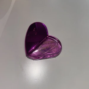 Lila hjärtformad parfymflaska - En unik hjärtformad parfymflaska i en djup lila nyans. Flaskan har en elegant design med en gyllene spraytopp. Perfekt för den som älskar en kombination av stil och funktion.