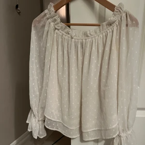 Vit offshoulder blus - Charmig vit offshoulder blus med långa ärmar och ett diskret prickigt mönster. Blusen har en luftig passform och elastiska kanter vid axlarna för en bekväm och stilren look.