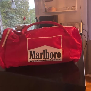 Röd sportväska från Marlboro - Säljer en cool röd sportväska från Marlboro World Championship Team. Väskan har ett stort huvudfack med dragkedja och bekväma bärhandtag. Perfekt för gymmet eller resan!