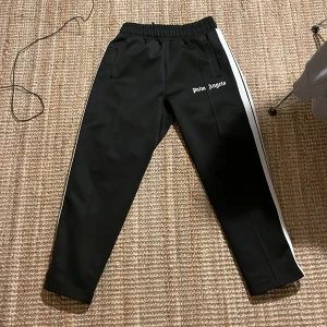 Svarta byxor från Palm Angels - Snygga svarta byxor från Palm Angels med vita ränder längs sidorna. De har en elastisk midja och en avslappnad passform, perfekt för en trendig look. Passar mig som är 176cm 63 kg