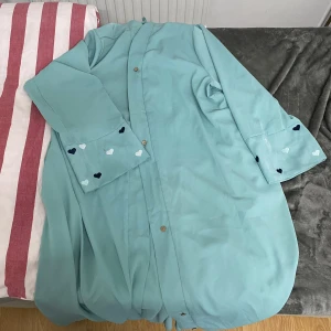 Turkos abaya med hjärtdetaljer - Säljer en turkos abaya med knappar och söta hjärtdetaljer på ärmarna. Perfekt för att ge en färgklick till din garderob. Kappan har en avslappnad passform och är långärmad.