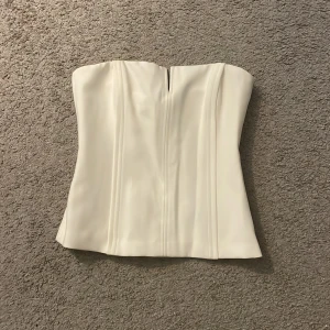 Vit korsett från Zara - Elegant vit korsett från Zara med dragkedja på sidan. Perfekt för en stilren look. Den är axelbandslös och har en smickrande passform. Prislappen är kvar och har ej använts. Köpts för 399kr. Varan säljs inte längre i Zara.